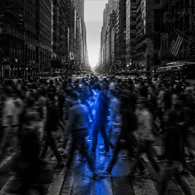 A solitary young man in a hoodie stands out in striking blue illumination, absorbed in his glowing phone amid a blurred crowd of pedestrians rushing through a bustling city crosswalk. The image powerfully illustrates the post's message, symbolizing how an individual can find clarity and signal amid the overwhelming noise and motion of modern life by tuning out distractions and focusing inward.