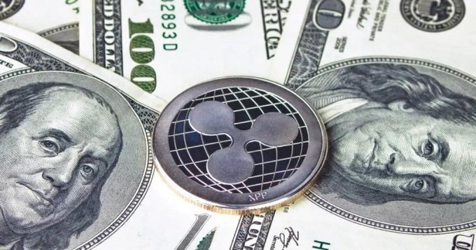 A physical XRP (Ripple) coin rests atop a fan of U.S. $100 bills featuring the portraits of Benjamin Franklin and Andrew Jackson, visually linking the cryptocurrency to traditional fiat currency. The composition reinforces the post's claim about central banks pegging XRP's value, while highlighting the $100 serial number (2893 D) visible on the bill in the background.