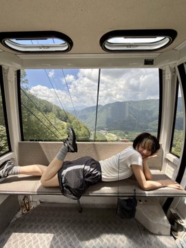 A young woman lounges in a cable car with a scenic mountain view in the background, enjoying a moment of peace before "catching her own lunch" in the countryside.