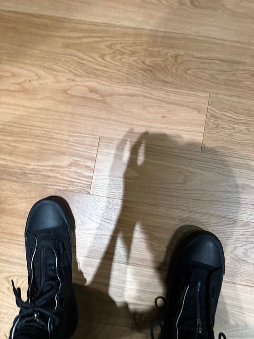 A shadow of a person holding up a peace sign appears on a wooden floor between their black sneakers, capturing a casual "clocking out" moment. The relaxed pose and hashtag "#LEO #리오" suggest a playful end-of-day vibe, with the peace sign symbolizing a carefree sign-off.