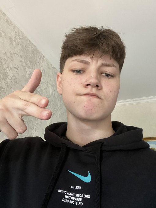 A young man wearing a black Nike hoodie points his finger toward the camera in a selfie taken indoors against a patterned wallpaper background.