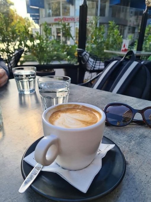 A cappuccino with latte art sits on a saucer at an outdoor café table, accompanied by a glass of water and sunglasses, with "onlybuys.c" visible on a storefront in the background.

The image creates a quiet, reflective mood that mirrors the contemplative tone of the post—suggesting a moment of pause to think clearly about what's substantive versus what's noise, much like the calm ritual of coffee paired with introspection about the project's unchanged core values.