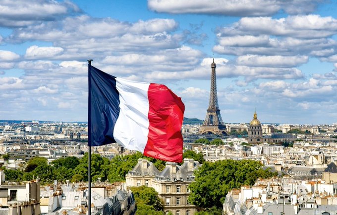 A French flag waving in the foreground with the Eiffel Tower and Parisian cityscape visible in the background under a partly cloudy blue sky.