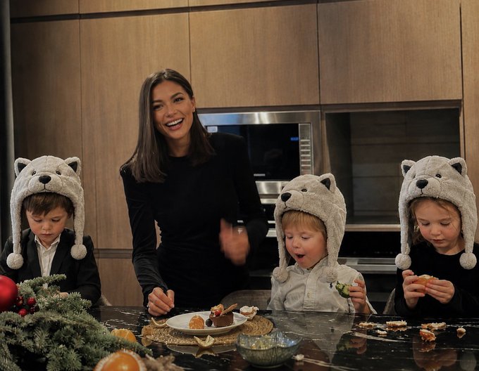 A laughing woman stands in a modern kitchen with three young children wearing matching gray wolf/dog trapper hats, gathered around a counter of cookies and treats — illustrating the joke about how the family's "wealth" came from the kids' dad simply howling "awoo." The cozy, festive scene with pine branches and holiday treats ties into the playful caption about their sudden riches from one silly sound.
