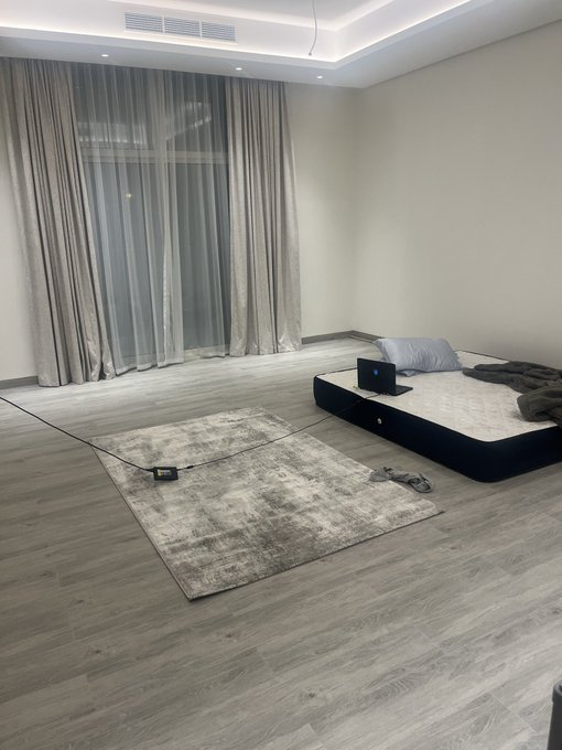 A bare mattress sits on a low frame in an otherwise empty room with gray curtains and a worn rug, illustrating the caption's joke about a makeshift sleeping setup lacking proper bedding.