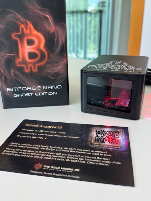 The image features a black box with a red neon "B" logo sitting next to a small card that has a QR code on it, which appears to be an invitation or information about Bitcoin mining. The card also includes a message encouraging people to support miners by purchasing their Bitcoins.