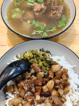 A meal consisting of a bowl of rice topped with braised pork and pickled greens, alongside a bowl of beef soup garnished with cilantro.