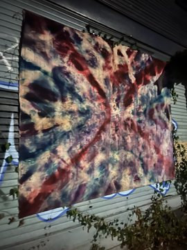 A tie-dye fabric tapestry or flag hanging outdoors against a house exterior, featuring a vibrant pattern in dark red, navy blue, cream, and tan colors arranged in a radial/spiral design.
