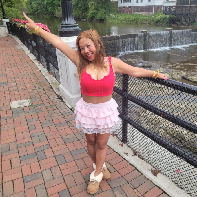 A smiling woman wearing a pink crop top and a pink ruffled skirt poses with her arms spread out on a brick walkway beside a river and a small waterfall.