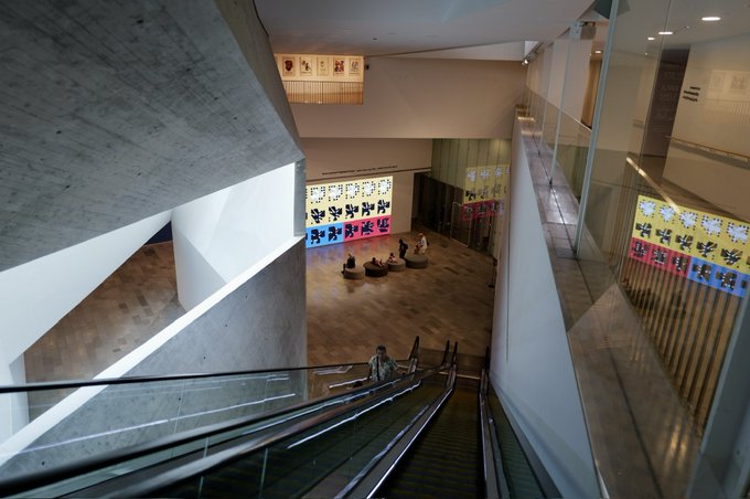An interior view looking down an escalator into a modern, multi-level museum or gallery space with concrete architecture, where visitors are viewing an art exhibit on the lower floor.