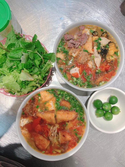 The image features two bowls of food, one containing soup with green vegetables and another bowl filled with fish. The bowls are placed on a table, along with some fresh greens such as lettuce and broccoli. There are also three small bowls of fruit, including grapes, scattered around the scene. The overall setting appears to be a casual dining experience or a meal shared between friends or family members.