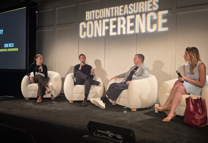Four panelists sit in white chairs on a stage at the 'BITCOINTREASURIES CONFERENCE,' with a smaller sign mentioning 'OAN HEID' and 'BITCOIN' visible to the side.