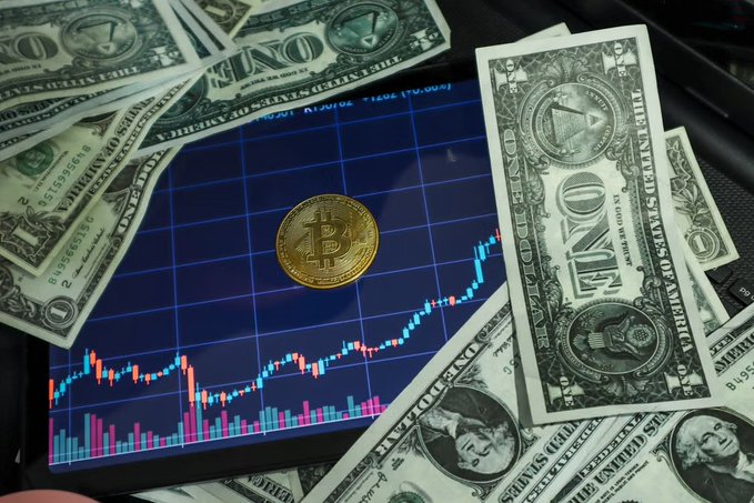 A tablet displaying a Bitcoin price chart with a physical Bitcoin token placed on the screen, surrounded by scattered US one-dollar bills.