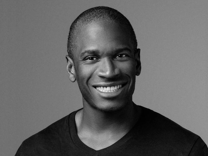 A black and white professional headshot portrait of a smiling bald man wearing a dark v-neck t-shirt against a plain grey background.