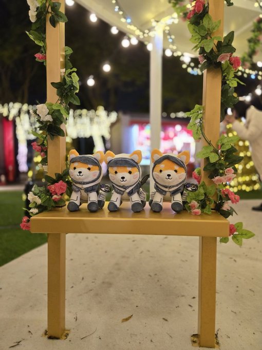 Three plush toys dressed as Shiba Inu samurai characters sitting on a yellow chair decorated with pink flowers and string lights at what appears to be a night event.