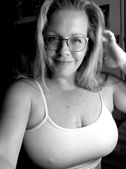 A black-and-white selfie of a smiling woman with blonde hair and glasses, wearing a light-colored tank top, with her hand touching her hair.