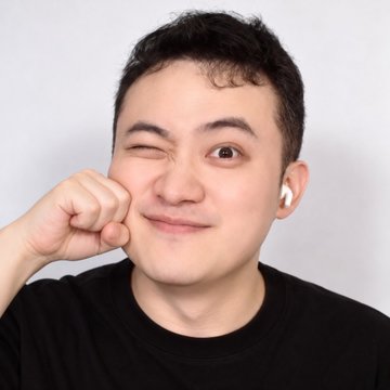 A man wearing a black shirt makes a playful face, winking one eye while pulling down his cheek with his fist, and wearing a white wireless earbud.