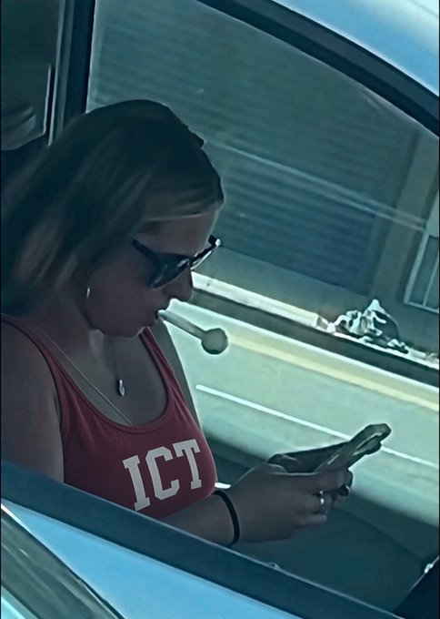 A woman wearing sunglasses and a red tank top with 'ICT' printed on it looks down at her phone while sitting in a car.