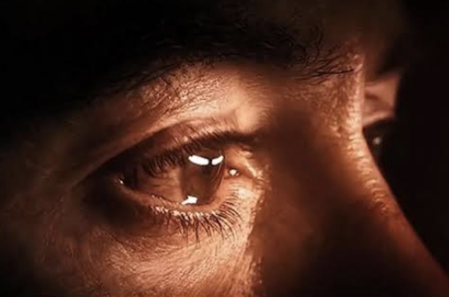 A close-up, dramatic shot of a person's eye reflecting light, with detailed visible eyelashes, skin texture, and warm lighting.