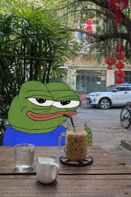 A Pepe the Frog cartoon character in a blue shirt drinks iced coffee at an outdoor wooden table with a small creamer pitcher, set against a backdrop of tropical plants, a building, red lanterns, and a parked white car.
