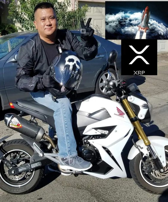 The image features a man sitting on a motorcycle, wearing a helmet that has "XRP" written on it. The man appears to be smiling for the camera while posing with his motorcycle. In the background of the photo, there are two cars parked nearby - one is black and the other is silver. Additionally, there's a rocket image in the top right corner of the photo.