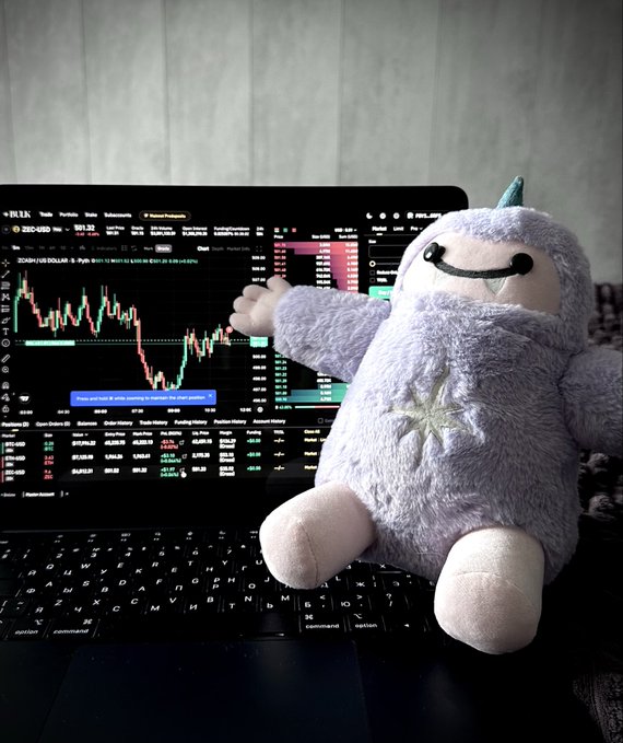 A small plush toy wearing a purple unicorn-like outfit sits on a laptop keyboard, with the screen displaying a trading chart on the RLK platform showing ZRO/USD with a price of $1.23.