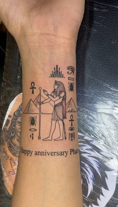 An Egyptian-themed tattoo on a person's wrist featuring an ankh, pyramids, a scarab beetle, and the text 'Happy anniversary Phar[...]'.
