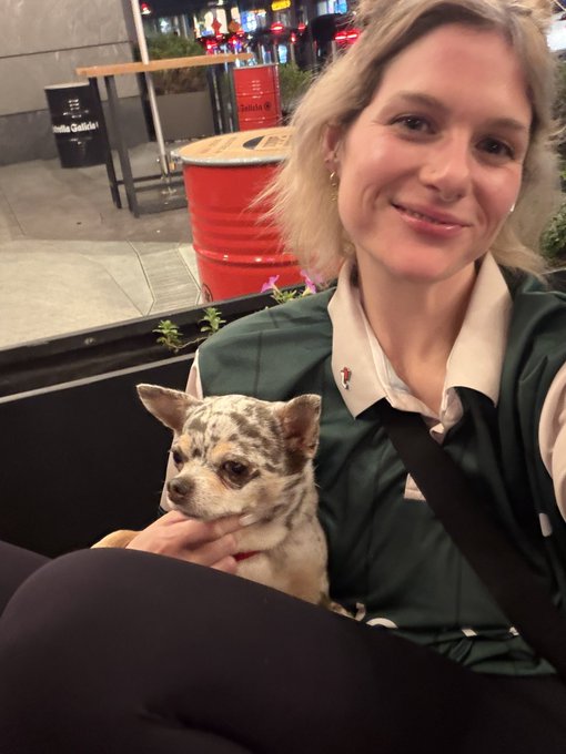 A blonde woman in a green jacket smiles at the camera while holding a small merle chihuahua, with red barrels and a bar visible in the background.