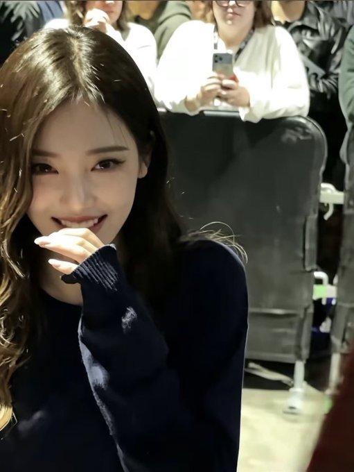 A close-up selfie shows a smiling woman with long wavy hair in a dark sweater inside a crowded venue, while people behind her hold up phones.