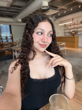 A woman with long curly hair takes a selfie in what appears to be a cafe or restaurant, wearing a black top and a bracelet.