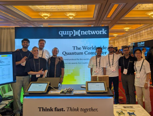 A group of eight people posing together at a trade show booth for 'QunaSys network' and 'QUAC', with signage promoting 'The Worldwide Quantum Computer' and the slogan 'Think fast. Think together.'