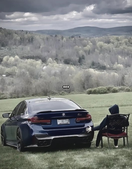 A person in a black hoodie sitting in a chair next to a dark blue BMW M5 parked on grass, overlooking a scenic forested valley under cloudy skies.