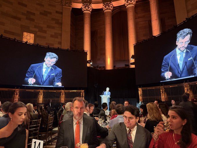 Formal gala event in an ornate hall with tall columns, featuring guests seated at tables while a speaker addresses the audience from a podium, with his image displayed on two large screens.