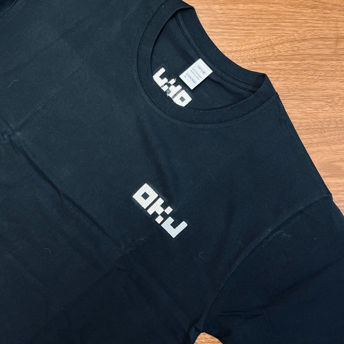 The image shows a black t-shirt with white text that reads "OKJ" printed on it, along with some other text. The t-shirt appears to be made of cotton or a similar fabric material. It is laid out flat on a wooden surface, and there are no visible objects in the background. The focus of the image seems to be on the t-shirt itself rather than any additional context or details about it.