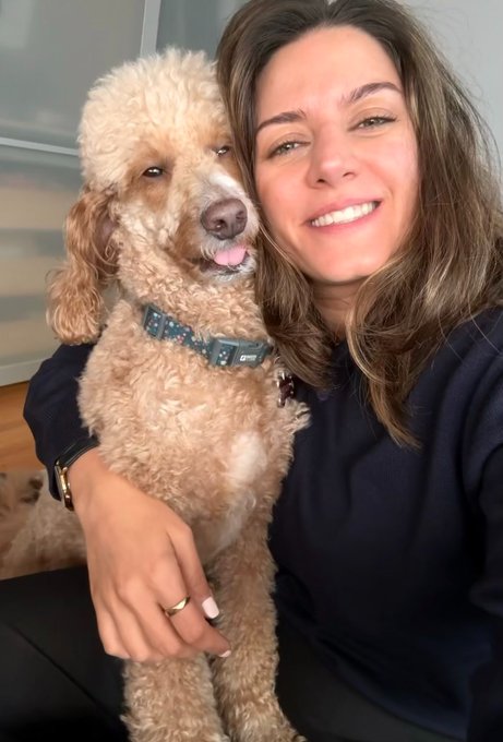 A smiling woman with brown hair poses for a selfie while hugging her fluffy apricot-colored dog, which wears a blue patterned collar.