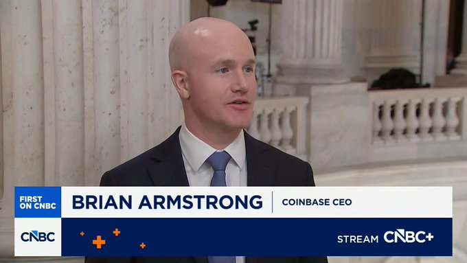 CNBC interview screenshot of Brian Armstrong, Coinbase CEO, in a suit and blue tie speaking in a stone-columned interior.