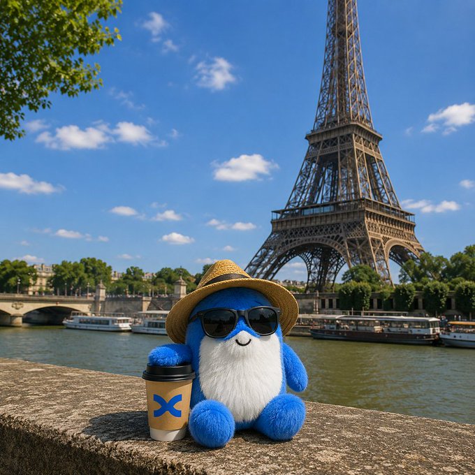 A blue plush character wearing a straw hat and sunglasses sits on a stone ledge holding a coffee cup with a blue "X" logo, posing in front of the Eiffel Tower along the Seine River in Paris—a charming travel moment greeting viewers with "hello."