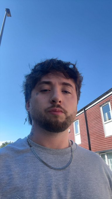 A selfie of a young man with curly brown hair and a short beard, wearing a gray t-shirt and a silver chain, standing outdoors against a clear blue sky.