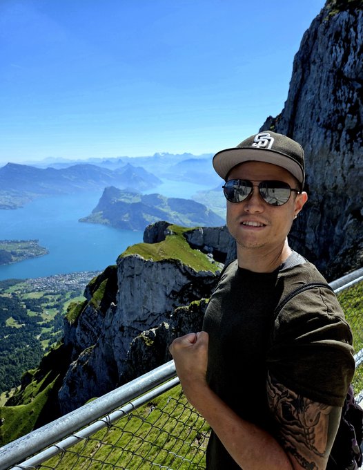 A man wearing a black "SF Giants" cap and sunglasses smiles triumphantly at the summit, with a stunning panoramic view of a lake and mountain range stretching out behind him — perfectly capturing the post's message that the toughest climbs lead to the best views, whether in the mountains or in chasing financial freedom through crypto.