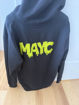 A black hoodie with "MAYC" displayed in bright yellow graffiti-style lettering on the back, representing Mutant Ape Yacht Club merchandise. The hoodie is photographed against a white wall on a wooden floor, matching the post's description of MAYC branded apparel being sold in size large.