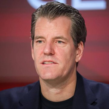 A man with short brown hair, wearing a dark blazer over a black shirt, speaks at what appears to be a conference with a red backdrop. The image likely illustrates the accompanying quote from Tyler Winklevoss predicting a 10X increase in Bitcoin's value, suggesting the man pictured may be the speaker being referenced.
