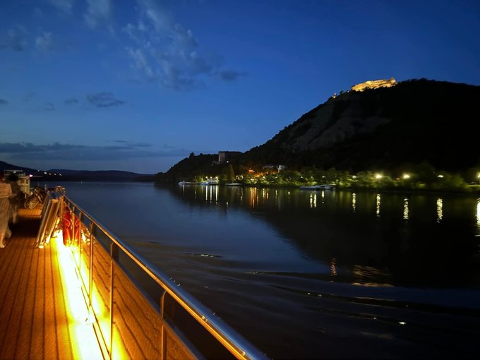 A nighttime view from a cruise ship's deck, looking across calm water toward a hillside with a brightly lit castle or fortress perched at the top.