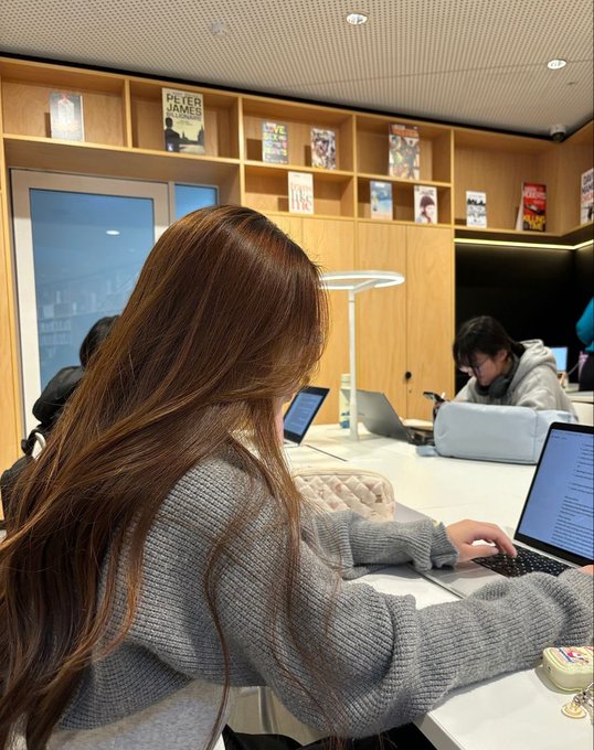 A photo of a young woman with long brown hair, viewed from behind, working on a laptop at a counter in what appears to be a bookstore, with shelves of books in the background.