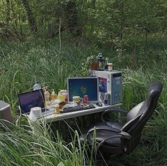 The image shows an outdoor workspace with two laptops, one of which has a keyboard attached to it. The laptop is placed next to a chair that is situated outside in a grassy area surrounded by trees. There are also several other items on the table, including a computer monitor and multiple cups scattered around. The scene seems to be set up for someone who prefers working outdoors or wants to take advantage of natural light while using their devices.