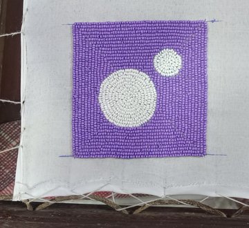 A close-up photograph of a handcrafted textile piece featuring purple and white beadwork or embroidery on a white fabric backing stretched over a wooden frame.