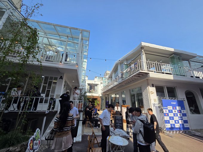 Outdoor plaza scene with people gathered around tables in front of modern buildings, one labeled 'B', under a clear blue sky with string lights.