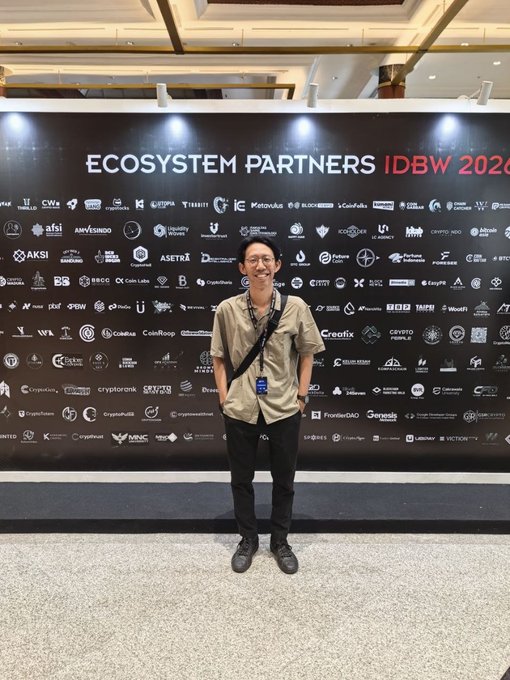 A young man wearing a tan short-sleeved shirt, black pants, and a lanyard stands smiling in front of a black backdrop featuring numerous cryptocurrency and Web3 company logos under the heading 'ECOSYSTEM PARTNERS IDBW 2024'.