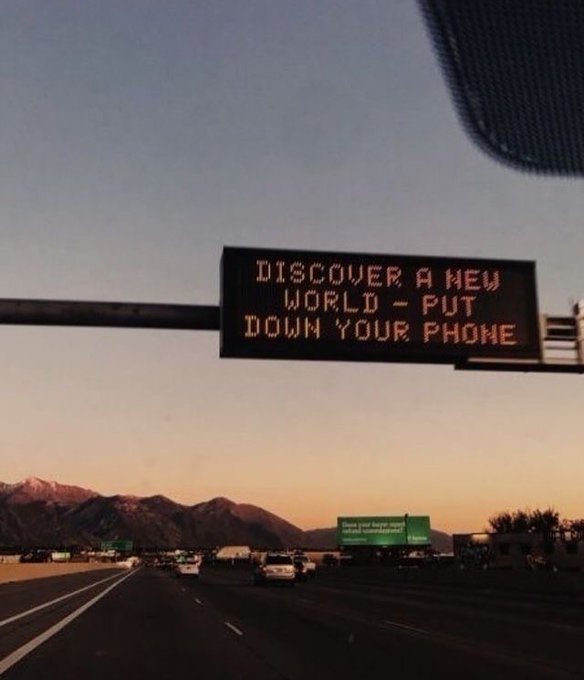 An electronic highway sign displays the message "DISCOVER A NEW WORLD - PUT DOWN YOUR PHONE" over a multi-lane freeway at sunset, with mountains visible in the background.