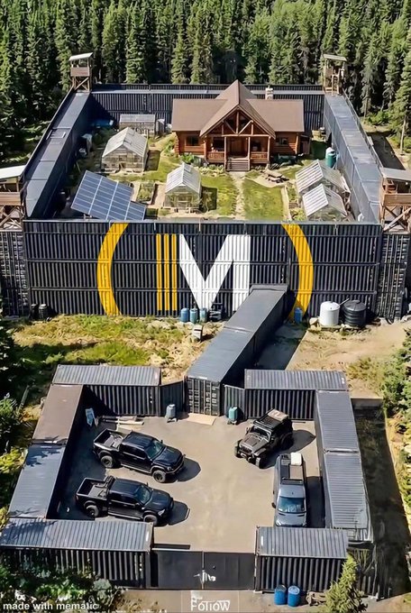 An aerial view of a fortified compound in a forested area constructed from shipping containers, featuring a large 'MW' logo painted on the wall, watchtowers at the corners, a central wooden lodge, greenhouses, solar panels, and several vehicles parked in a gated area.