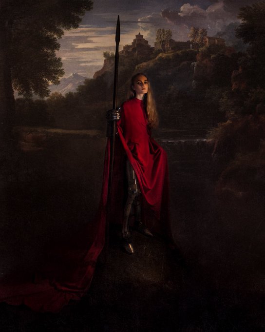A solitary female figure in flowing red robes stands holding a long spear in a moody, romantic landscape with distant mountains and a hilltop fortress, capturing the artistic theme of feminine strength and legacy referenced in the post.